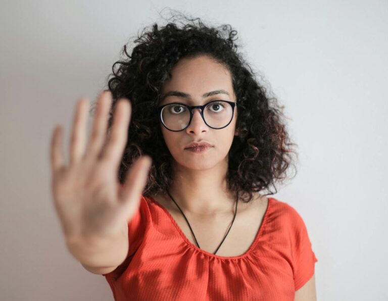 portrait photo of woman in red top wearing black framed eyeglasses holding out her hand in stop gesture